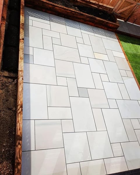 Patio with white rectangular tiles arranged in a herringbone pattern, bordered by wooden beams and green grass