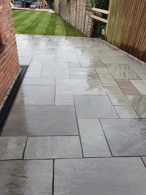 Wet stone patio with large rectangular pavers leading to a manicured lawn and garden fence