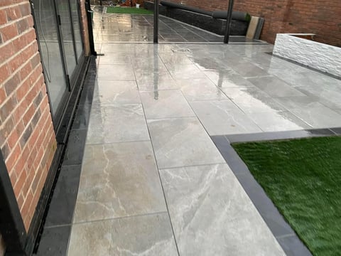 Modern patio with large gray slate tiles, dark edging, artificial turf, and brick wall alongside