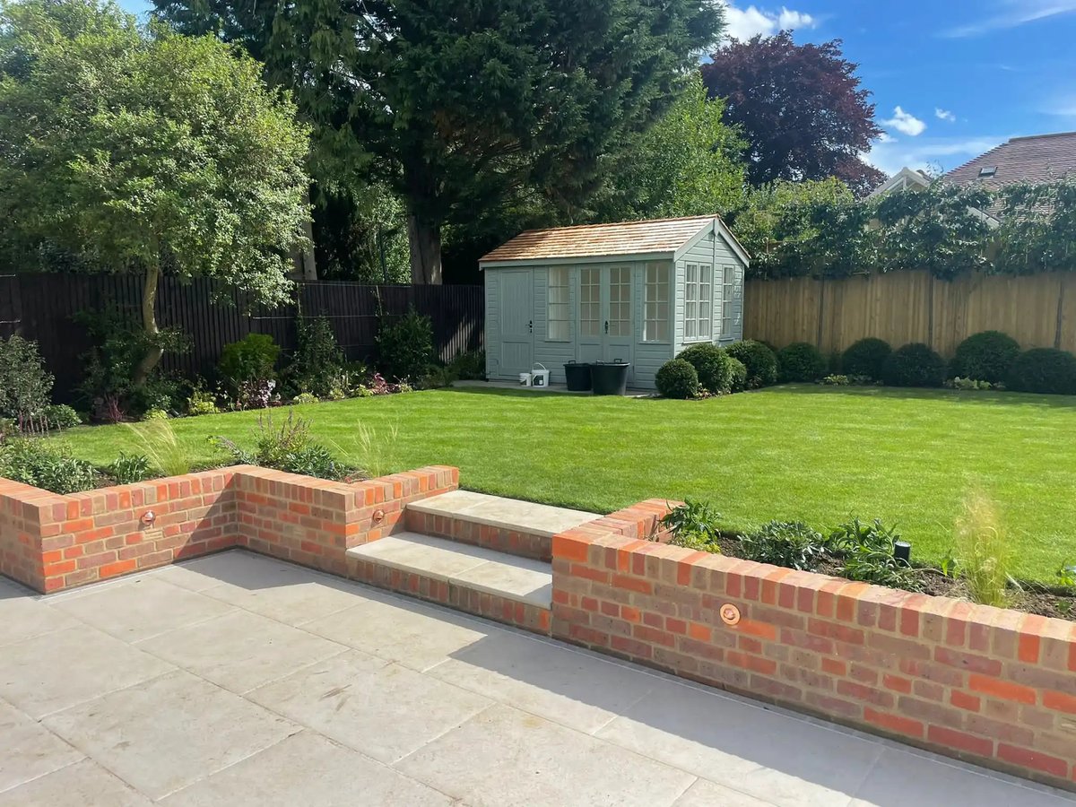Backyard garden with brick raised beds, paved patio, green lawn, shed, and mature trees under blue sky