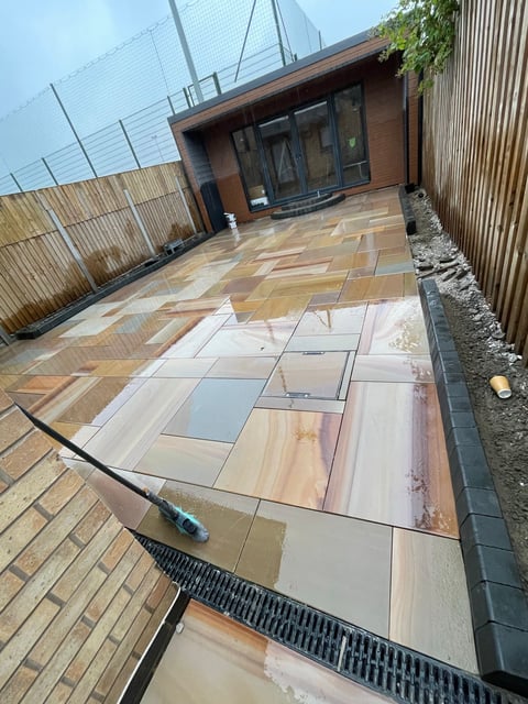 Modern patio with mixed stone and wood paving being cleaned with pressure washer, brick building and wooden fence in background