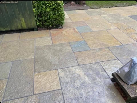 Patio with mixed-color stone pavers in geometric pattern, green fence and shrub visible in background