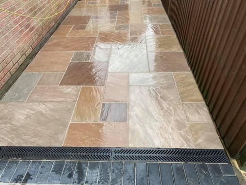 Outdoor patio with varied earth-tone stone tiles in mixed pattern, bordered by dark edging strip, adjacent to corrugated metal wall